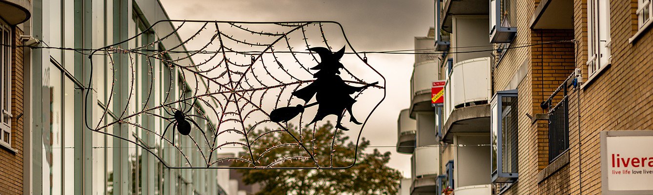 Een spinnenweb met een spin en een heks op een bezem hangt als lichtdecoratie tussen de gevels boven de winkelstraat.