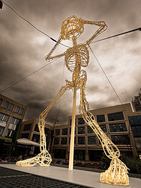 Een meters hoog skelet van verlicht draadwerk staat met wijd gespreide benen op de bestrating, de ene arm omhoog langs het hoofd en de andere zijwaarts gestrekt, met achter zich de gevels van het Stadshart en een donkere wolkenlucht.